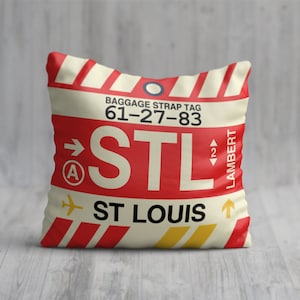 May include: A red and white throw pillow with a luggage tag design. The tag says "Baggage Strap Tag 61-27-83" and "STL" with an arrow pointing to the right. Below the tag is the text "ST LOUIS" with an arrow pointing up. The tag also says "LAMBERT" on the right side.