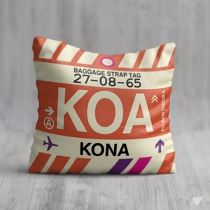 May include: A cream-coloured pillow with a red and white striped design. The pillow features a luggage tag with the text "BAGGAGE STRAP TAG 27-08-65 KOA KONA" and an arrow pointing to the right. The text "ELLISON ONIZUKA" is on the right side of the pillow.
