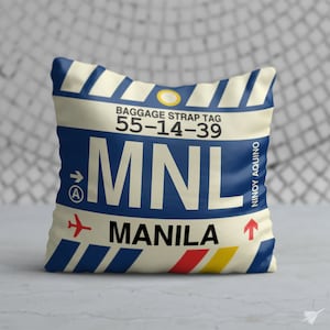 May include: A white pillow with a blue and white striped design. The pillow has a luggage tag design with the text "BAGGAGE STRAP TAG 55-14-39 MNL MANILA" and the airport code "MNL" in large blue letters. The text "NINOY AQUINO" is printed vertically on the right side of the pillow.