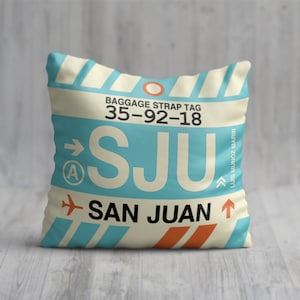 May include: A white cushion with a blue and orange luggage tag design. The tag says "BAGGAGE STRAP TAG 35-92-18" and "SJU SAN JUAN".