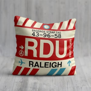 May include: A white and red throw pillow with a luggage tag design. The tag says "RDU Raleigh" and has the baggage strap tag number "43-96-58".