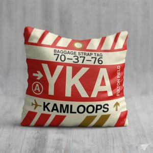 May include: A white pillow with red and gold stripes features a luggage tag with the text "BAGGAGE STRAP TAG 70-37-76 YKA FULTON FIELD KAMLOOPS" and a red arrow pointing to the right with a letter "A" inside a circle.