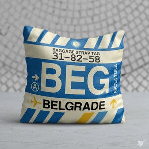 May include: A white pillow with a blue and yellow luggage tag design. The tag says "BEG" and "BELGRADE" with the airport code "31-82-58" and "NIKOLA TESLA" written on the side.