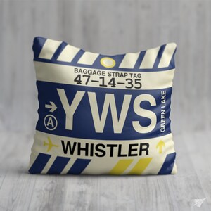 May include: A cream-coloured pillow with a blue and yellow luggage tag design. The tag reads "YWS Whistler" with the airport code "47-14-35" and the words "Baggage Strap Tag" and "Green Lake".