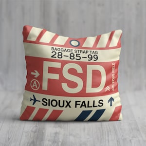 May include: A cream-colored pillow with red and blue stripes. The pillow has a luggage tag design with the text "BAGGAGE STRAP TAG 28-85-99 FSD SIOUX FALLS JOE FOSS FIELD".