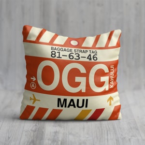 May include: Square pillow with a luggage tag design. The pillow features the text "BAGGAGE STRAP TAG 81-63-46 OGG MAUI KAHULUI" in orange and white on a cream background. Includes an airplane graphic and arrow.