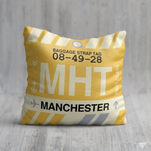 May include: A square cushion with a yellow background and a design resembling a baggage strap tag. The text "MHT" is prominently displayed, along with "Manchester" and other details. The cushion has a first-class label and a striped pattern.