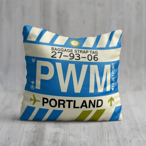 May include: A white pillow with a blue and yellow luggage tag design. The tag says "BAGGAGE STRAP TAG 27-93-06 PWM PORTLAND" with an airplane icon.