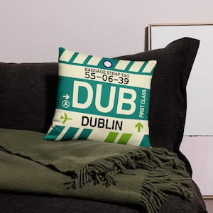 May include: A white and green pillow with a luggage tag design. The tag says "BAGGAGE STRAP TAG 55-06-39 DUB DUBLIN" with an airplane icon and an arrow pointing to the right. The tag also says "FIRST CLASS" with an arrow pointing up.