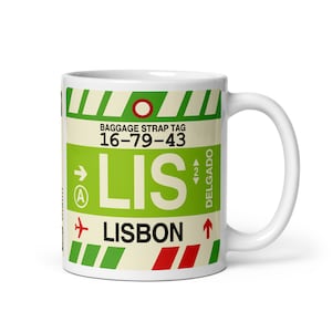 May include: White ceramic mug with a design resembling a baggage tag in green, red, and white. The text "LIS LISBON" and "DELGADO" is visible. The mug has a curved handle.