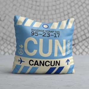 May include: A blue and white cushion with a luggage tag design. The tag reads "BAGGAGE STRAP TAG 95-23-17 CUN CAN CUN", with an arrow pointing to the right and a small aeroplane icon. The word "TURISTA" is written vertically on the right side of the tag.