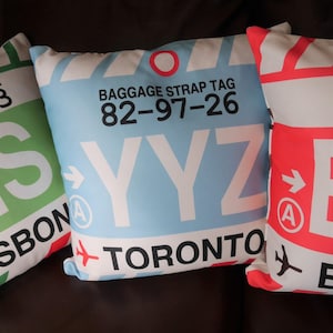 May include: Three decorative throw pillows with a luggage tag design. The pillows are white with red, blue, and green accents. The text on the pillows reads "Baggage Strap Tag 82-97-26 YYZ TORONTO" and "LISBON".