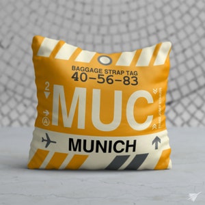 May include: A yellow and white pillow with a luggage tag design. The tag says "Baggage Strap Tag 40-56-83 MUC Munich Franz Josef Strauss".