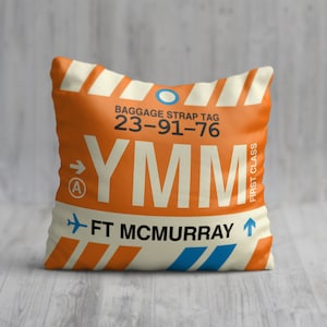 May include: Orange and white cushion with a luggage tag design. The tag has the airport code YMM, the city "FT MCMURRAY", and the baggage strap tag number 23-91-76. The tag also has the words "FIRST CLASS" and an arrow pointing to the right.