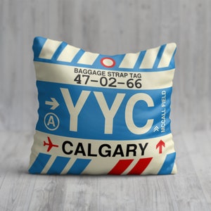 May include: A white and blue throw pillow with a luggage tag design. The tag says "YYC CALGARY" with the baggage strap tag number "47-02-66" and "MCCALL FIELD".
