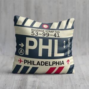 May include: A cream-colored pillow with a blue and red luggage tag design. The tag has the text "BAGGAGE STRAP TAG 53-99-41" and "PHL PHILADELPHIA" with a red airplane icon. The tag also has a blue circle with a white "A" and a red arrow pointing right.