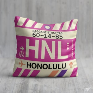 May include: A pink and white throw pillow with a luggage tag design. The tag says "HNL" for Honolulu, Daniel K. Inouye International Airport, and "Baggage Strap Tag 60-14-85".