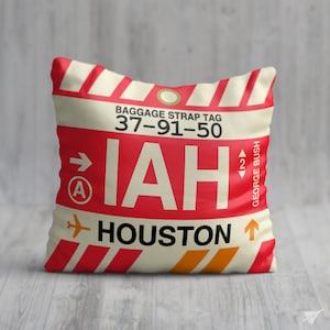 May include: A white and red pillow with a luggage tag design. The tag says "Baggage Strap Tag 37-91-50 IAH HOUSTON" with an arrow pointing to the right and a small "A" in a circle. The tag also has an arrow pointing up with the text "GEORGE BUSH" next to it.
