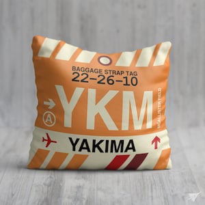 May include: Orange and white throw pillow with a luggage tag design. The tag has the airport code YKM and the city name YAKIMA. The text "BAGGAGE STRAP TAG 22-26-10" is printed on the tag. The tag also has the words "MCALLISTER FIELD" printed on the side.