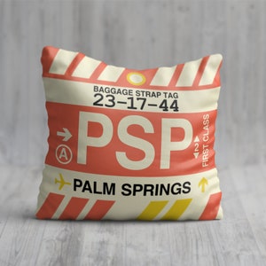 May include: A red and white pillow with a luggage tag design. The tag says "BAGGAGE STRAP TAG 23-17-44 PSP PALM SPRINGS" and has a yellow airplane icon. The pillow is decorated with yellow and red stripes.