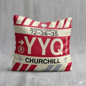 May include: A white and red pillow with a luggage tag design. The tag says "Baggage Strap Tag 72-36-35 YYQ CHURCHILL" and has a red arrow pointing to the right with a letter "A" inside a circle. The tag also has a small airplane icon and an arrow pointing up. The text "FIRST CLASS" is printed vertically on the right side of the tag.