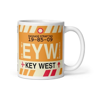 May include: White ceramic mug featuring a design inspired by a baggage strap tag. The design includes the text "EYW" and "KEY WEST" in a bold font, along with the numbers "19-85-09" and other graphic elements in orange, red, and white.