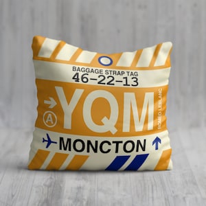 May include: A white pillow with a yellow and orange luggage tag design. The tag has the airport code "YQM" and the city "MONCTON" printed on it. The tag also has the text "BAGGAGE STRAP TAG 46-22-13" printed on it.