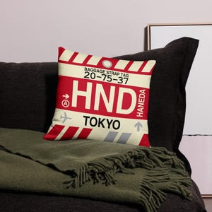 May include: A red and white pillow with a luggage tag design. The tag says "Baggage Strap Tag 20-75-37 HND Haneda Tokyo".