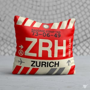 May include: Square cushion with a red background and a luggage tag design. The cushion features the text "ZRH" in large white letters, along with "ZURICH" and other details. The design includes white and red stripes and the words "FIRST CLASS".
