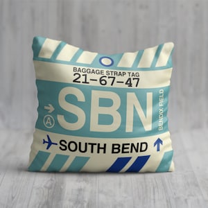 May include: A cream-colored pillow with a blue and white luggage tag design. The tag reads "BAGGAGE STRAP TAG 21-67-47 SBN SOUTH BEND" with an arrow pointing to the right and a small airplane icon. The tag also says "BENDIX FIELD" on the right side.