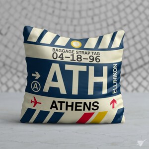 May include: A white cushion with a blue and white striped design. The cushion features a luggage tag design with the text "ATH" and "ATHENS" in large blue letters. The luggage tag also includes the text "BAGGAGE STRAP TAG 04-18-96" and "ELLINIKON" in smaller letters.