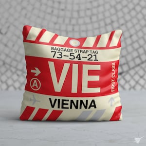 May include: A cream-coloured pillow with red and grey stripes. The pillow has a luggage tag design with the text "BAGGAGE STRAP TAG 73-54-21 VIE VIENNA FIRST CLASS" printed on it.