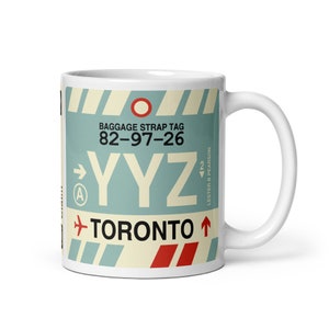 Peut inclure: Mug en céramique blanche avec un graphique d'une étiquette à bagages pour l'aéroport international Pearson de Toronto (YYZ). L'étiquette est bleue, jaune et rouge avec le texte "BAGGAGE STRAP TAG 82-97-26 YYZ TORONTO" et une flèche pointant vers la droite.