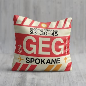 May include: Square cushion with a luggage tag design. The cushion features a red and cream colour scheme with the text "GEG SPOKANE" and "BAGGAGE STRAP TAG 93-30-45". The design includes an airplane graphic and arrows.