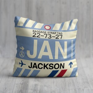May include: A square pillow with a light blue background and a design resembling a baggage strap tag. The text "JAN JACKSON" is prominently displayed, along with the numbers "22-73-27" and the name "MEDGAR EVERS". The pillow has a cream-colored border.
