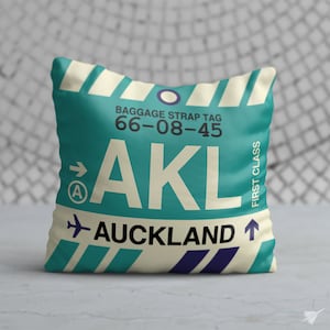 May include: A white pillow with a blue and green luggage tag design. The tag says "AKL AUCKLAND" and "BAGGAGE STRAP TAG 66-08-45" with an arrow pointing to the right and a small "A" in a circle. The tag also says "FIRST CLASS" with an arrow pointing up.