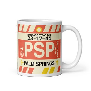 May include: White ceramic mug featuring a red and white luggage tag design. The tag reads "Baggage Strap Tag 23-17-44 PSP Palm Springs" with a yellow aeroplane icon.
