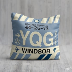 May include: A blue and white cushion with a luggage tag design. The tag reads "Baggage Strap Tag 44-26-73 YQG Windsor" with an arrow pointing to the right and a small "A" in a circle. The tag also says "First Class" on the right side.