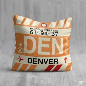 May include: A white pillow with an orange and red luggage tag design. The tag says "DENVER" and "BAGGAGE STRAP TAG 61-94-37" with an arrow pointing to the right and the word "STAPLETON" on the right side.