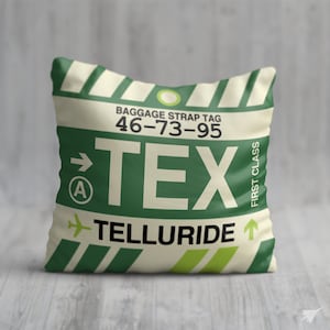 May include: A white pillow with green stripes and a luggage tag design. The tag says "BAGGAGE STRAP TAG 46-73-95 TEX TELLURIDE" with a green arrow pointing right and a green airplane icon.