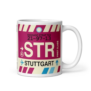 May include: White ceramic mug with a design resembling a baggage strap tag. The design features the letters "STR", the word "Stuttgart", and other text in maroon, cream, and blue. The mug has a curved handle.