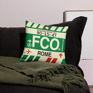 May include: A decorative pillow with a green and white baggage strap tag design. The pillow features the text "FCO ROME" in large white letters, with a red airplane icon and an upward arrow. The pillow is on a dark gray couch.