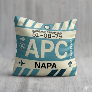 May include: A decorative cushion with a vintage luggage tag design. The tag features the text "BAGGAGE STRAP TAG 51-08-79", "APC", "NAPA", and "FIRST CLASS".