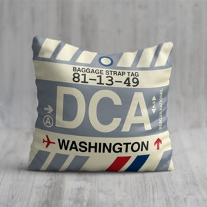May include: A cream-coloured pillow with a blue and white luggage tag design. The tag has the text "Baggage Strap Tag 81-13-49 DCA WASHINGTON" and a small aeroplane icon. The tag also has a blue circle and a red arrow pointing upwards.