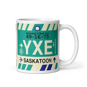 May include: White ceramic mug featuring a blue, green and white airport baggage tag design. The tag reads "Baggage Strap Tag 49-16-73 YXE Saskatoon".
