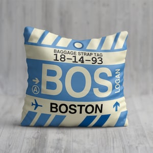 May include: A cream-coloured pillow with a blue luggage tag design. The tag has the text "BAGGAGE STRAP TAG 18-14-93 BOS LOGAN BOSTON" and an arrow pointing to the right with the letter "A" inside a circle.