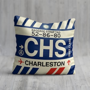 May include: A white pillow with a blue and red luggage tag design. The tag says "Baggage Strap Tag 52-86-80 CHS CHARLESTON" and "First Class".