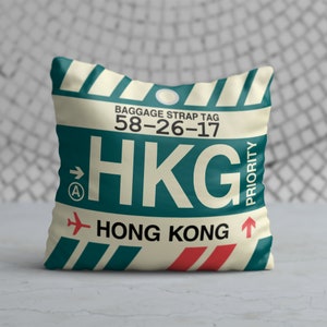 May include: A cream-coloured pillow with a blue and red luggage tag design. The tag says "BAGGAGE STRAP TAG 58-26-17 HKG HONG KONG" with an arrow pointing to the right and a priority label.