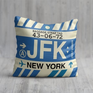 May include: Cushion featuring a blue and cream baggage strap tag design. The text includes "JFK", "NEW YORK", and "JOHN F KENNEDY". The pillow has a vintage aesthetic, with a design reminiscent of travel.