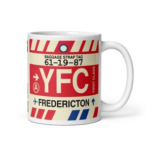 Puede incluir: Taza de cerámica blanca con un diseño de etiqueta de equipaje de aeropuerto rojo, blanco y azul. La etiqueta dice "Baggage Strap Tag 61-19-87 YFC FREDERICTON" con un círculo azul y una flecha.